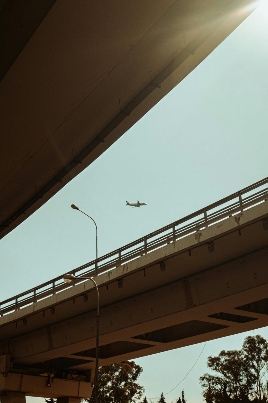 Photograph of an airplane flying above a city bridge captured in sunlight, showcasing modern infrastructure and travel.