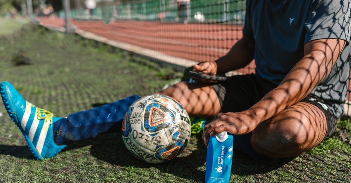 A soccer player takes a break, sitting on a grassy field with a soccer ball and water bottle.
