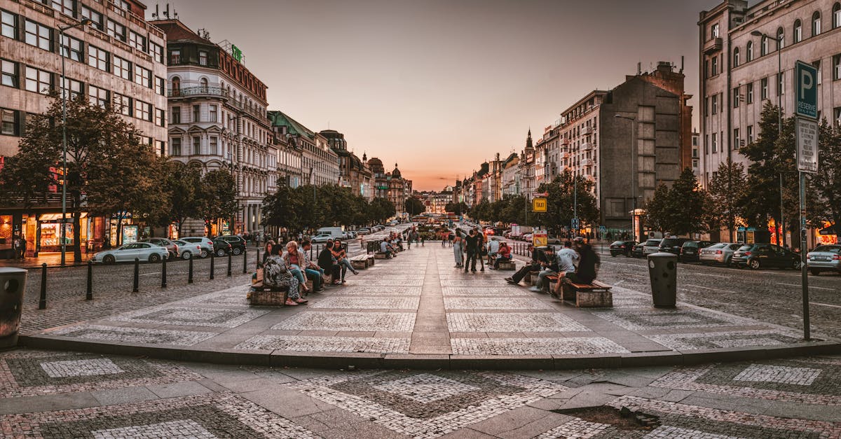 A bustling urban street in Prague at sunset, featuring people, cars, and vibrant architecture.